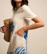 Vestido corto de viscolino color marfil con escote redondo, mangas cortas y bolsillos laterales. Cuenta con un estampado frontal en tonos de azul que representa un paisaje de palmeras y olas del mar.
