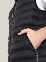Chaleco acolchado de nailon color negro, con cuello alto, cierre frontal de cremallera con detalles distintivos de la marca y bolsillos laterales con cierre. Presenta el logo de Tommy Hilfiger bordado en el pecho.