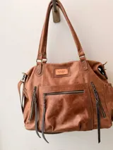 Bolso maternal de cuero marrón con doble asa de hombro, correa ajustable, múltiples bolsillos frontales y laterales con cierre, y detalles de tiras trenzadas.
