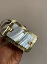 Cinturón de cuero negro con hebilla rectangular de nácar con marco dorado.