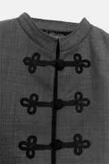 Blazer de corte cropped confeccionado en tejido de lana gris, con cuello redondo y cierre frontal mediante alamares decorativos. Presenta bolsillos delanteros con solapa y puños a contraste en color negro.