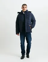 Campera acolchada de color gris, con capucha desmontable, cierre frontal y bolsillos laterales con cremallera. Presenta un diseño de costuras horizontales y detalles en color naranja en los tiradores de los cierres.