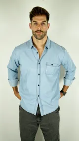 Camisa de jean de manga larga en tono azul claro, con cuello clásico, cierre frontal de botones oscuros y un bolsillo en el pecho.
