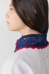 Camisa de lino blanco con cuello y puños tejidos a mano en crochet de color azul con borde rojo. Presenta cierre frontal con botones, bolsillo lateral y costuras a la vista en contraste.