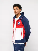 Campera deportiva Umbro del Club Nacional de Football, modelo Summit. Presenta un diseño con bloques de color en azul, rojo y blanco, con capucha, cierre frontal y bolsillos laterales con cierre. Incluye el logo de la marca y el escudo del club en el pecho.