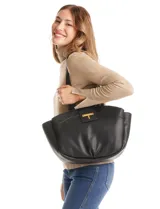 Cartera tote negra de ecocuero con base redondeada y pliegues decorativos. Cuenta con una solapa central con herraje dorado en forma de "T" y asas dobles para hombro y mano. El interior tiene triple compartimento.