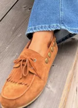Náuticos de gamuza color suela con flecos en la parte delantera y detalle de tachas metálicas circulares en el lateral. Diseño artesanal con cordones frontales y suela plana.