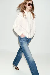 Camisa blanca de cuello redondo con volantes, manga larga con puños elásticos y cierre frontal con botones forrados. Presenta detalles de puntilla y jaretas en el delantero.