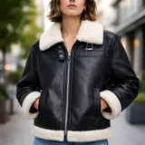 Campera tipo aviadora de ecocuero negro con forro interior, cuello, puños y borde inferior de corderito blanco. Presenta cierre frontal metálico, dos bolsillos laterales y detalles de hebillas metálicas en el cuello.