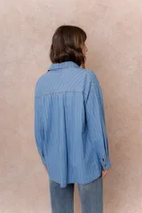 Camisa oversize de tela liviana con efecto denim, color azul con rayas verticales blancas. Presenta cuello camisero, cierre frontal con botones, bolsillo aplicado en el pecho, hombros caídos y mangas largas con puños abotonados.