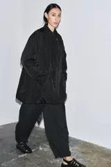 Chaqueta oversize de nylon en color negro, con cuello redondo subido, manga larga y cierre frontal con botones a presión ocultos. Presenta bolsillos delanteros y forro interior a tono.