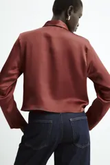 Blusa de tejido satinado con cuello solapa, manga larga y detalle de abertura drapeada en el pecho. Cierre frontal con botón forrado.