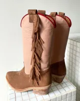 Botas estilo tejano de caña alta, confeccionadas en cuero vacuno. Presentan una combinación de colores en beige y marrón texturizado, con detalle de flecos laterales y borde superior en color rojo.
