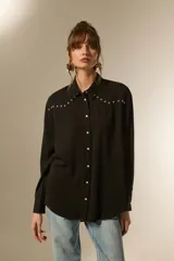 Camisa negra de viscosa con corte holgado, cuello clásico, cierre frontal de botones y detalle de tachas metálicas en la línea de los hombros.