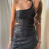 Vestido corto asimétrico con un hombro descubierto y dos breteles finitos que se atan cruzados en la espalda. El vestido es de lentejuelas plateadas y tiene un corte en la cintura.