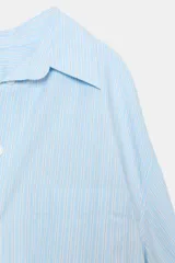 Camisa de corte oversize con estampado de rayas verticales en tonos celeste y blanco. Presenta cuello solapa, escote en pico, manga larga con puños abotonados y un bolsillo de plastrón en el pecho.