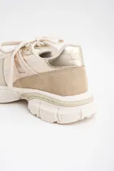 Zapatilla deportiva de diseño urbano en tonos neutros, combinando paneles de malla, gamuza y detalles metalizados en color dorado. Cuenta con suela robusta y acordonado frontal.