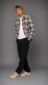 Camisa tipo overshirt de corte holgado, confeccionada en tejido de textura suave con estampado de cuadros en tonos beige, negro y azul. Presenta cuello clásico, cierre frontal con botones y dos bolsillos delanteros con solapa en el pecho.