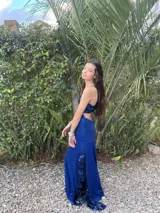Vestido largo de satén color marrón con acabado metalizado, escote en V, tirantes finos cruzados en la espalda y diseño con corte bajo el busto y volados en la falda.