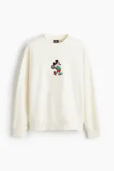 Buzo de tejido de algodón color crema, corte oversized y cuello redondo. Presenta un pequeño bordado central de Mickey Mouse vestido con ropa deportiva y sosteniendo una raqueta de tenis.