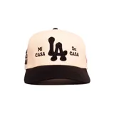 Gorra con visera plana de color negro y corona en tono natural. Presenta bordados frontales en color negro con el monograma central y las frases "Mi Casa" y "Su Casa" a los lados.