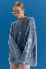 Sweater de tejido de punto grueso en color azul grisáceo, con escote redondo y diseño holgado. Presenta estampado gráfico en el frente y en una de las mangas, con terminaciones acanaladas en cuello, puños y ruedo.