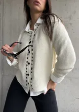 Cardigan de punto color natural con botones negros y detalle de bordado lineal en contraste a lo largo de la botonera. Presenta cuello redondo, mangas largas con puños acanalados y ajuste holgado.