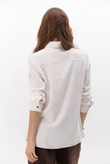 Camisa beige de manga larga, con cuello clásico, botones marrones y dos bolsillos frontales. Presenta costuras decorativas en contraste.