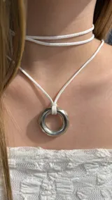 Gargantilla tipo choker compuesta por un cordón blanco de gamuza sintética que rodea el cuello y un dije central en forma de aro metálico facetado.
