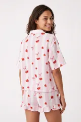 Pijama compuesto por una camisa de manga corta con solapas y botones frontales, y un short con detalles de volados en las piernas. Presenta un estampado de rayas verticales rosadas y blancas con motivos de cerezas.