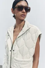 Chaleco gris acolchado con cuello alto, cierre frontal con cremallera metálica, bolsillos de plastrón en delantero y bajo ajustable con cordones.