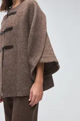 Capa de lana fina sin teñir, con diseño de corte amplio y mangas tipo kimono. Presenta cierre frontal mediante tres alamares de cuero en tono marrón.