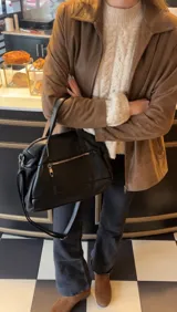 Cartera de mano y hombro color beige, de material símil cuero, con forma estructurada y redondeada. Posee dos asas cortas superiores y una correa larga ajustable y desmontable. Cuenta con un bolsillo exterior con cierre en la parte frontal.