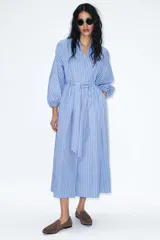 Vestido midi de corte camisero con estampado de rayas verticales en tonos azul, blanco y rojo. Presenta cuello redondo con escote en pico, mangas largas abullonadas con puños elásticos y cinturón ajustable del mismo tejido. Incluye bolsillos laterales ocultos y cierre frontal cruzado con botones.