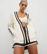 Cardigan deportivo de corte oversized, color blanco con detalles en marrón oscuro. Presenta escote en V, cierre frontal con botones a presión, mangas largas con franjas laterales y puños acanalados. Incluye un parche circular con letras en el pecho.