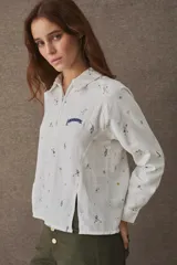 Remera estilo polo de manga larga, confeccionada en una mezcla de algodón y lino. Presenta un cierre frontal parcial, cuello camisero y aberturas laterales en el frente. El diseño incluye un estampado integral con motivos deportivos y un detalle bordado en el pecho.