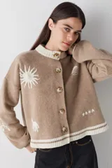 Cardigan de lana color crema con cuello redondo, cierre frontal de botones y mangas largas con puños ajustados. Presenta bordados decorativos en el frente que incluyen un sol, olas y estrellas, con detalles en azul en el cuello y el ruedo.