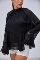 Blusa negra de cuello alto con volados en el pecho y mangas largas con transparencias. Presenta un diseño escalonado con volados en la parte inferior.