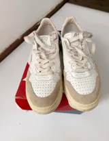 Championes Autry de diseño retro, color blanco con detalles en gamuza beige y suela de tono tostado, con cordones claros y logo lateral de la marca con la bandera de Estados Unidos.