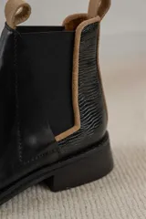Botas tipo Chelsea de cuero negro, con paneles laterales elásticos y detalle de textura grabada en el talón. Presentan un borde en color beige y tiradores en la parte superior.
