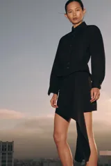 Falda corta de tiro medio, color negro, con detalle de pliegues y volante asimétrico en el delantero. Cierre con cremallera oculta en la costura.