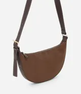 Cartera bandolera de formato media luna, confeccionada en material sintético efecto gamuza color marrón. Presenta correa larga ajustable con costuras a la vista y cierre superior con cremallera.