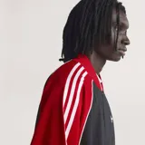 Campera deportiva Adidas SST de corte holgado, confeccionada en tejido de malla color negro con mangas rojas y detalles de franjas blancas características de la marca. Cuenta con cuello tipo bomber, cierre frontal completo y logo del trifolio bordado en el pecho.