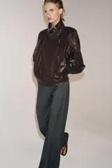 Chaqueta de corte amplio estilo biker en tejido efecto piel de color verde oscuro, con cuello alto envolvente, cierre frontal asimétrico de cremallera metálica, bolsillos ocultos en costura y forro interior.