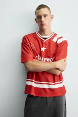 Camiseta de manga corta color rojo, corte boxy fit, con cuello pico ribeteado en blanco. Presenta un estampado frontal con el texto "Social Leagues", una estrella blanca y un escudo con el número "4". Tiene detalles de franjas blancas en el bajo y en los hombros.