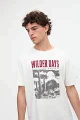 Remera de manga corta en color blanco crudo, confeccionada en jersey de algodón pesado de tacto suave. Presenta un calce relaxed y un diseño gráfico estampado en el frente con la leyenda "WILDER DAYS" en rojo y un paisaje desértico en tonos grises.