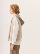 Chaleco de punto con capucha, confeccionado en lana. Presenta un diseño bicolor con cuerpo en tono beige de textura acanalada y capucha en tono marrón. Incluye una etiqueta de cuero en la parte posterior.