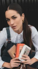 Billetera mediana con cierre perimetral, confeccionada en cuero sintético rosa y detalle central de tejido tipo telar con rayas verticales en amarillo, rosa y negro. Incluye correa de mano desmontable y placa metálica con logo de la marca.