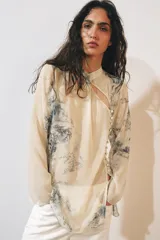 Camisa de cuello redondo y manga larga con diseño de estampado artístico en tonos azules sobre fondo beige. Presenta un cierre frontal asimétrico con botones forrados a tono y una abertura lateral en el escote.