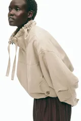 Campera beige en mezcla de lino, con cuello elevado con cordón ajustable, cierre frontal y bolsillos laterales con solapa y botón. Presenta hombros caídos y elástico revestido en el bajo.